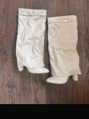 Beige slouchy stövlar med klack - Säljer ett par beige slouchy stövlar med bred klack och rundad tå. Stövlarna har en dekorativ rem med metallspänne upptill och är gjorda i ett mjukt mockaliknande material. Perfekta för dig som vill ha en trendig och avslappnad look.