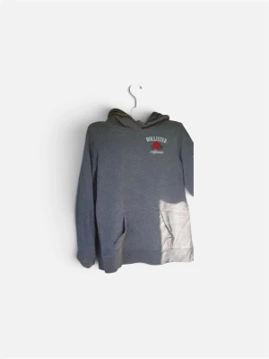 Grå hoodie från Hollister med ros - Snygg grå hoodie från Hollister med en röd ros och texten 'california' tryckt på bröstet. Hoodien har huva och en stor magficka framtill. Perfekt för en avslappnad och trendig look. Tillverkad i mjukt material för extra komfort.