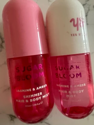 Sugar Bloom Jasmine & Amber Mist - Två flaskor Sugar Bloom Jasmine & Amber Hair & Body Mist från Yes Studio. Båda har rundad form och rosa toner, en med rosa lock och en med vitt lock. Doften är en mix av jasmin och amber, och ena flaskan har shimmer-effekt. Perfekt för att fräscha upp hår och kropp.