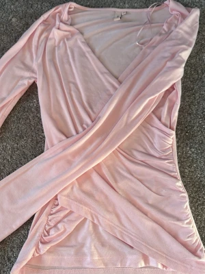 Ljusrosa omlottopp från Nelly - Supersnygg ljusrosa omlottopp från Nelly i mjukt och stretchigt material. Toppen har lång ärm, v-ringning och smickrande omlottdesign som ger en feminin siluett. Perfekt att styla med jeans eller kjol för en trendig look.