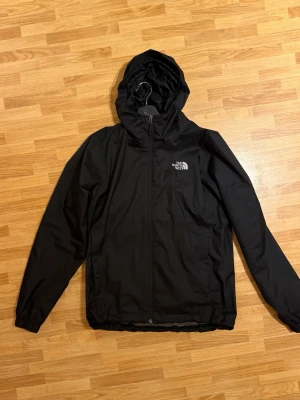 The North Face windbreaker  - Snygg svart regnjacka från The North Face med  justerbar huva och dragkedja framtill. Jackan har två sidofickor och justerbar nederkant. Tillverkad i ett vattenavvisande material som passar perfekt för regniga dagar. Logotypen syns tydligt på bröstet… knappt använd, passar mig hyfsat men vill ha annat är 180.