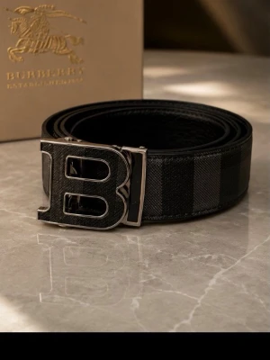 Svart Burberry bälte med B-spänne - Snyggt svart bälte från Burberry med diskret rutigt mönster och ett stort B-format metallspänne. Bältet har en exklusiv känsla och passar perfekt till jeans eller kostymbyxor. Materialet är skinn med detaljer i metall.