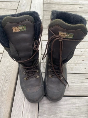 Chiruca Hunter varmfodrade kängor - Mörkbruna höga kängor från Chiruca, modell Hunter, med grov sula och varm, svart fleecefodring. Kängorna har metallhakar för snörning och slitstark ovandel i läder och textil. Perfekta för kalla dagar och utomhusaktiviteter.
