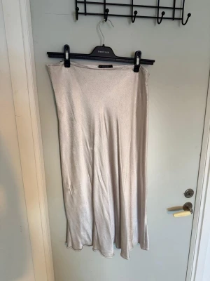 Ljusbeige långkjol i satin - Säljer en ljusbeige långkjol i mjukt satinliknande material. Kjolen har en enkel och stilren design med lätt glansig yta och faller snyggt längs benen. Perfekt att matcha med en crop top eller stickad tröja.