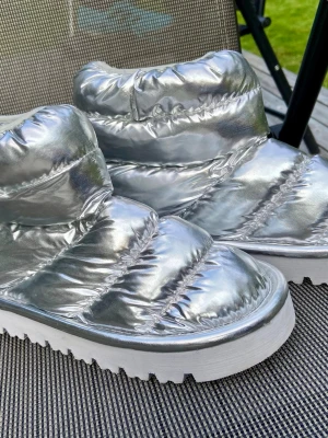 Silvriga moon boots med platåsula stl.38 H&M - Säljer ett par riktigt coola moon boots i metallic-silver med tjock, vadderad ovandel och vit platåsula. Skorna har en quiltad look och ger en futuristisk vibe. Perfekta för dig som vill sticka ut i vinter och hålla fötterna varma. Sparsamt använda endast 2 gånger.