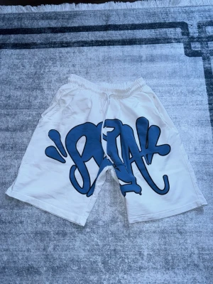 Vita shorts med blått graffiti-tryck - Säljer ett par vita shorts med stort blått graffiti-tryck framtill. De har elastisk midja med snörning och en bakficka. Perfekta för en avslappnad streetwear-look.