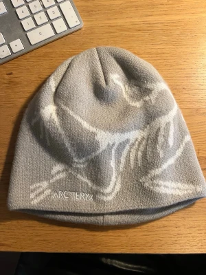 Ljusgrå mössa från Arc'teryx - Snygg ljusgrå mössa från Arc'teryx med vitt grafiskt mönster och broderad logga framtill. Tillverkad i mjukt material som sitter skönt på huvudet. Perfekt för kalla dagar och outdoor-äventyr.