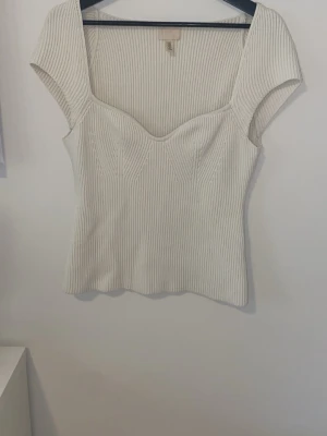 Vit ribbad topp från H&M - Säljer en vit ribbad topp från H&M med markerad byst och breda axelband. Toppen har en figurnära passform och är tillverkad i ett stretchigt material som ger en snygg siluett. Perfekt att styla med jeans eller kjol.