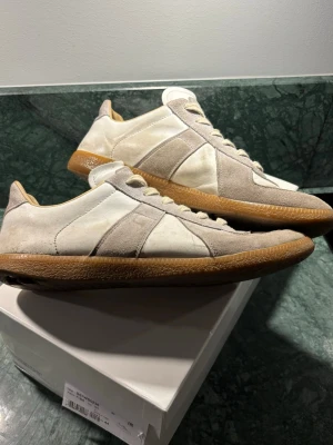 Maison margiela Gats - Säljer dessa Margiela Gats som troligen sett sina bästa dagar men har nog fortfarande mycket kvar att ge, snygga sneakers i vitt och beige med ovandel i mocka och läder. Skorna har en klassisk låg siluett och bruna gummisulor som ger en retrovibe. Tyvärr hål i sulorna. Dustbag och låda följer med. Hör gärna av dig för fler bilder eller frågor!🤝