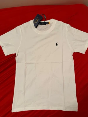 Vit t-shirt från Polo Ralph Lauren - Klassisk vit t-shirt från Polo Ralph Lauren med korta ärmar och rund hals. T-shirten har den ikoniska broderade loggan på bröstet och är tillverkad i mjuk bomull för en skön känsla. Perfekt basplagg till alla outfits.
