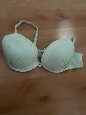Ljusgrön push-up bh med spetsdetaljer - Snygg ljusgrön push-up bh med vadderade kupor och spetsdetaljer längs kanten. Axelbanden är justerbara och bh:n har en liten dekorativ rosett framtill. Stängs med hyskor bak. Materialet känns mjukt och bekvämt. Finns lite svarta små prickar på ena kupan. 