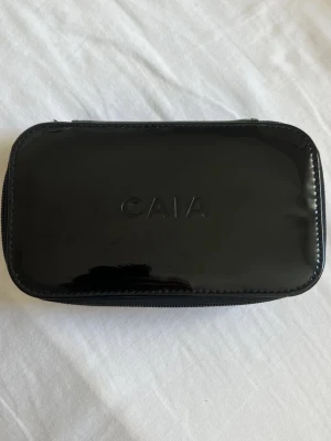 CAIA Svart necessär  - Stilren svart necessär från CAIA i glansigt syntetmaterial. Rektangulär form med dragkedja runtom och diskret CAIA-logga på locket. Perfekt för smink eller småsaker när du är på språng.
