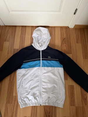 Lacoste vit och marinblå vindjacka - Snygg vindjacka från Lacoste i vitt, marinblått och ljusblått. Jackan har huva, dragkedja framtill och två fickor. Klassisk krokodillogga på bröstet och elastiska muddar vid ärmar och midja. Perfekt nu till hösten för en sportig och stilren look. 