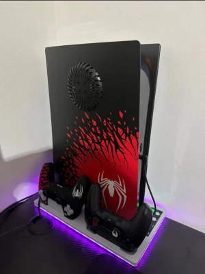 Playstation 5 Spider-Man Edition - Playstation 5 konsol med Spider-Man design i svart och rött, med vita spindelmotiv. Konsolen står på en laddstation med LED-belysning och två matchande handkontroller. Snygg och unik look för dig som gillar superhjältar och gaming.