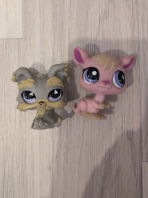 Littlest Pet Shop figurer hund & kängru - Två söta Littlest Pet Shop figurer, en grå hund med beige detaljer och stora ögon samt en rosa kängru med beige detaljer och blå ögon. Perfekta till samlingen eller som leksaker. Båda figurerna har klassiska LPS-stilen med stora huvuden och uttrycksfulla ansikten.