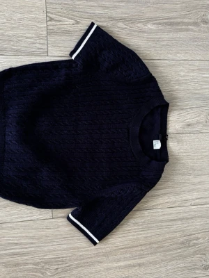 Mörkblå stickad t-shirt med vita ränder - Snygg mörkblå stickad t-shirt med korta ärmar och vita ränder vid ärmslut och nederkant. Tröjan har ett diskret kabelstickat mönster framtill och ribbad halsringning. Perfekt för en stilren och avslappnad look.