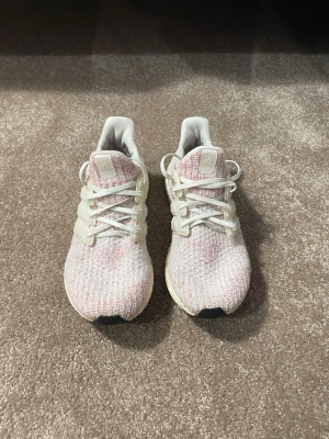 Vita och rosa Adidas sneakers - Säljer ett par snygga Adidas sneakers i vitt med inslag av ljusrosa och grått. Skorna har stickad ovandel, vita snören och klassiska tre ränder på sidan. Sulan är vit med svart yttersula och ger en sportig vibe. Perfekta för dig som gillar stilrena och bekväma sneakers.