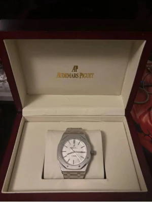 Audemars Piguet Royal Oak silverklocka - Exklusiv Audemars Piguet Royal Oak klocka i silver med ikonisk åttakantig boett och länkarmband i metall. Urtavlan är ljus med ett rutigt mönster och tydliga indexmarkeringar. En riktigt stilren accessoar för dig som gillar lyx och klassisk design. Klockan är automatisk ur och är vattentät