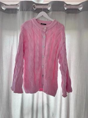 Rosa kabelstickad kofta Gina Tricot - Supersöt rosa kabelstickad kofta från Gina Tricot med rund halsringning och vita knappar framtill. Koftan har ribbade muddar vid ärmslut och nederkant, och är gjord i ett mjukt stickat material. Perfekt för lager på lager och en poppig look.