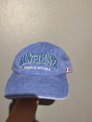 Ljusblå keps Saint-Tropez - Snygg ljusblå keps med broderad text 'SAINT-TROPEZ FRENCH RIVIERA' framtill och liten fransk flagga på sidan. Klassisk böjd skärm och justerbar passform bak. Perfekt accessoar för en avslappnad och trendig look.