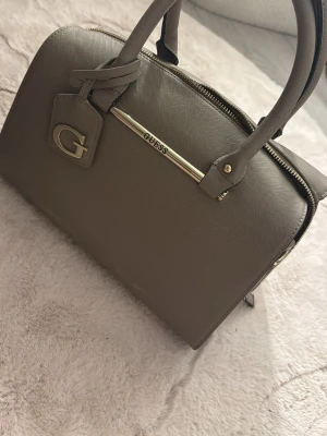 Taupe handväska från Guess - Snygg taupefärgad handväska från Guess i strukturerat konstläder. Väskan har två handtag, guldfärgade detaljer och en dekorativ tag med stort G. Klassisk boxig form och dragkedja upptill.