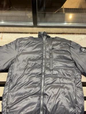 Svart dunjacka med bröstfick från Canada goose - Snygg svart dunjacka med hög krage och dragkedja framtill. Jackan har en praktisk bröstficka med dragkedja och en patch på ärmen. Perfekt för kalla vinterdagar tack vare det värmande dunet och den stilrena designen.