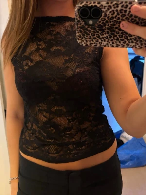 Svart spetsig ärmlös topp - Säljer en svart, ärmlös topp i spets med blommigt mönster. Toppen är croppad och har en transparent look som ger en edgy vibe. Perfekt att styla med höga byxor eller kjol för en trendig outfit.