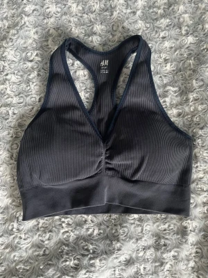 Mörkblå sport-bh från H&M - Säljer en mörkblå sport-bh från H&M med ribbad struktur och bred resår under bysten. Bh:n har brottarrygg och är tillverkad i ett stretchigt material som ger bra stöd. Perfekt för träning eller en aktiv vardag.
