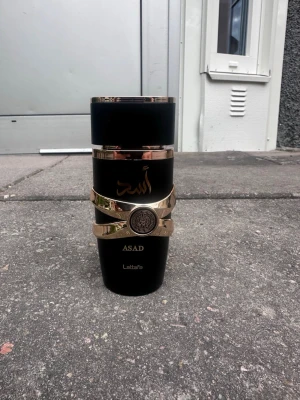Lattafa Asad parfym svart/guld - Lattafa Asad parfym i en elegant svart flaska med gulddetaljer och arabiska inskriptioner. Flaskan har en unik form med guldfärgat band runt mitten och ett dekorativt emblem. Perfekt för dig som gillar exklusiv design och orientaliska dofter.