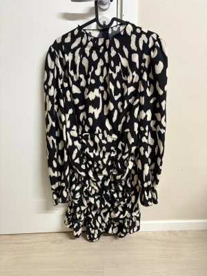 Svartvit kort klänning med leopardmönster - Kort klänning med svart botten och vita leopardfläckar. Klänningen har långa ärmar, rynkade detaljer framtill och volang vid nederkanten. Materialet känns som polyester och passformen är normal.