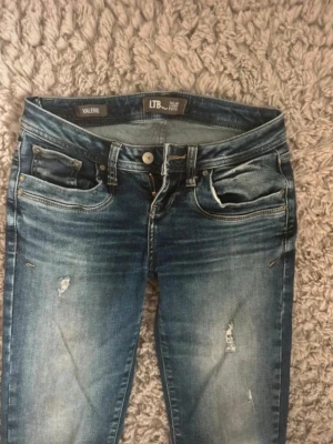 Low waisted jeans från LTB - Säljer ett par blå jeans från LTB i modellen VALERIE. Jeansen har en snygg tvätt med slitningar och små hål på benen, fem fickor och klassisk knappgylf. Perfekta för en avslappnad och trendig look.