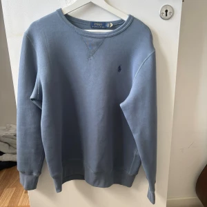 Blå sweatshirt från Polo Ralph Lauren - Klassisk blå sweatshirt från Polo Ralph Lauren med rund halsringning och diskret broderad logga på bröstet. Tröjan har ribbade muddar vid ärmslut och nederkant samt en avslappnad passform. Perfekt för en stilren och bekväm look.