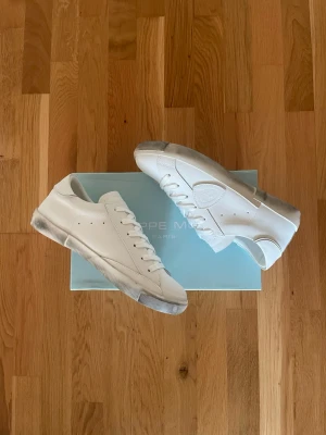 Vita låga sneakers från Philippe Model - Stilrena vita sneakers från Philippe Model i lågt snitt med klassisk siluett. Skorna har vita skosnören, diskreta håldetaljer på sidan och en vit sula med lätt slitning. Tillverkade i slätt läder för en clean och modern look.
