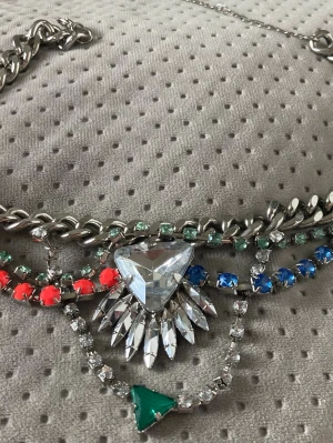 Färgglatt statementhalsband med stenar - Unikt statementhalsband i silverfärgad metall med stora, glittrande stenar i olika färger. Halsbandet har en mix av klara, blå, gröna och röda stenar samt en stor triangelformad kristall i mitten. Kedjan är grov och detaljerad med flera lager av gnistrande dekorationer.