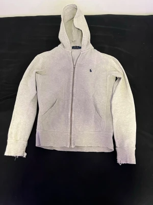 Grå zip hoodie från Polo Ralph Lauren - Snygg grå hoodie med dragkedja från Polo Ralph Lauren. Klassisk design med huva, två fickor fram och den ikoniska lilla loggan broderad på bröstet. Tillverkad i mjukt sweatshirtmaterial som är skönt att ha på sig. Perfekt för en avslappnad och stilren look.