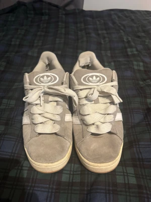 Adidas campus 00 - Snygga grå sneakers från Adidas i mjuk mocka med vita detaljer och klassiska tre ränder på sidan. Skorna har en låg profil, vita skosnören och Adidas-logga på plösen. Yttersulan är i gummi med beige ton.