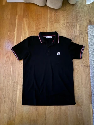 Svart moncler piké - Klassisk svart pikétröja med krage och knappar framtill. Tröjan har röda och vita detaljer på krage och ärmslut samt en liten logga på bröstet. Perfekt till jeans eller shorts för en clean look.