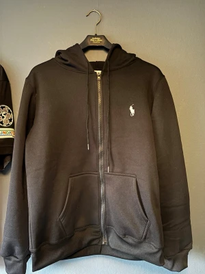 Svart zip hoodie från Polo Ralph Lauren - Snygg svart hoodie med dragkedja från Polo Ralph Lauren. Klassisk design med huva, två stora fickor framtill och den ikoniska vita logon broderad på bröstet. Tillverkad i mjukt material som passar perfekt till en avslappnad stil. Helt oanvänd säljes då den är för liten för mig. Storlek S