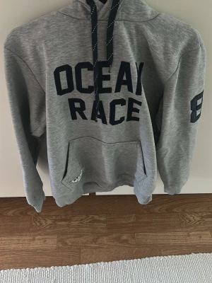 Grå hoodie med tryck OCEAN RACE - Snygg grå hoodie med svart text 'OCEAN RACE' på bröstet och siffran 8 på ärmen. Klassisk känguruficka framtill och justerbar huva med svarta snören. Tillverkad i mjukt sweatshirtmaterial, perfekt för en avslappnad stil.