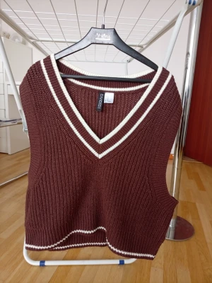 Brun stickad väst från H&M Divided - Snygg brun stickad väst från H&M Divided med vita ränder vid v-ringningen och nederkanten. Västen har en ribbad struktur och är perfekt att styla över en skjorta eller t-shirt för en trendig look.