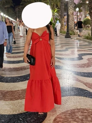 Röd långklänning med rosettdetalj - Snygg röd långklänning med axelbandslös design och stor rosettdetalj framtill. Klänningen har en liten cut-out under bysten och volang nedtill för extra flow. Perfekt för sommarkvällar och semester. Använt 1 gång, skriv för fler bilder 