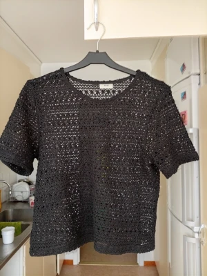 Svart virkad topp med kort ärm - Snygg svart virkad topp med korta ärmar och rund hals. Toppen har ett genombrutet mönster som ger en luftig och trendig look. Perfekt att styla över ett linne eller bikini. Passar dig som gillar en unik och bohemisk vibe.