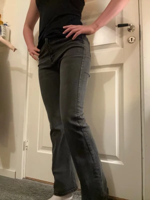 Svarta bootcut jeans med fyra fickor högmidjade - Säljer ett par svarta bootcut jeans med klassisk femficksdesign och dragkedja med knapp. Jeansen har lätt utsvängda ben och subtila slitningar framtill. Materialet är denim med en mjuk känsla och sömmar i matchande färg.