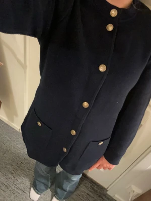Mörkblå cardigan från Hampton Republic - Mörkblå lång cardiganjacka från Hampton Republic med guldknappar framtill och på fickorna. Två stora fickor fram och rundad nederkant. Stickad i mjukt material och har lång ärm. Enkel och stilren design som passar till mycket.