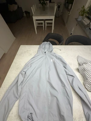 Ljusgrå vindjacka från Under Armour - Ljusgrå vindjacka med huva från Under Armour. Jackan har dragkedja framtill, två sidofickor och justerbar nederkant. Tillverkad i lätt och tunt material, perfekt för blåsiga dagar. Diskret logga på bröstet och enkel, sportig design.
