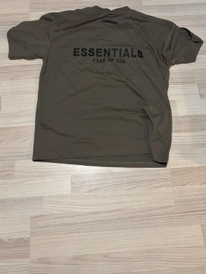 Olivgrön Essentials Fear of God tee - Säljer en olivgrön t-shirt från Fear of God Essentials med stor svart logga på ryggen. Oversized passform och klassisk rund hals. Perfekt för dig som gillar streetwear och vill ha en enkel men snygg tee med statement-tryck.