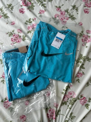 Turkost träningsset från Nike - Säljer ett fräscht turkost träningsset från Nike med t-shirt och shorts. Båda plaggen har den klassiska Nike-loggan i svart och silver. Materialet är lätt och andas, perfekt för träning eller sportaktiviteter. Shortsen har resårmidja och dragsko för skön passform.