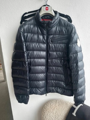Moncler Genius 1952 - Snygg svart pufferjacka med hög krage och quiltad design. Jackan har dragkedja framtill, två framfickor med dragkedja och en bröstficka. Materialet är glansigt och jackan har en patch på ena ärmen. Perfekt för kalla vinterdagar. Navy jacket. Size 3. Fits M/L. 100% authentic ✅