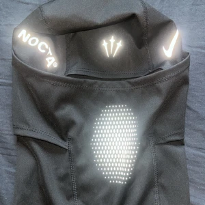 Svart balaclava från Nike NOCTA - Säljer en svart balaclava från Nike NOCTA med reflekterande detaljer och logotyper. Materialet är stretchigt och andas, perfekt för att täcka ansiktet. Snygg design med NOCTA- och Nike-logga samt prickigt mönster framtill.