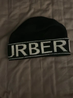 Svart mössa från Burberry - Snygg svart mössa från Burberry med vit logotyptext runt kanten. Klassisk och enkel design som passar till det mesta. Tillverkad i mjukt material som håller dig varm under kalla dagar.